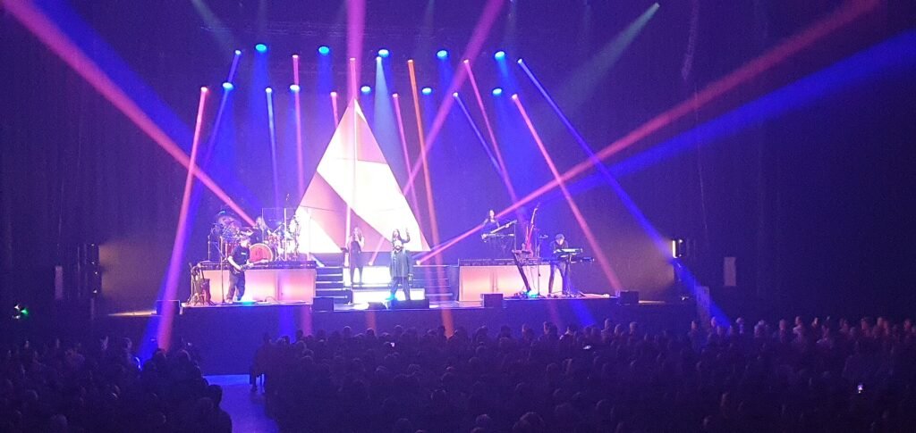 Alphaville on stage under pink and blue light beams in front of a large white triangle, seen from the back of an arena