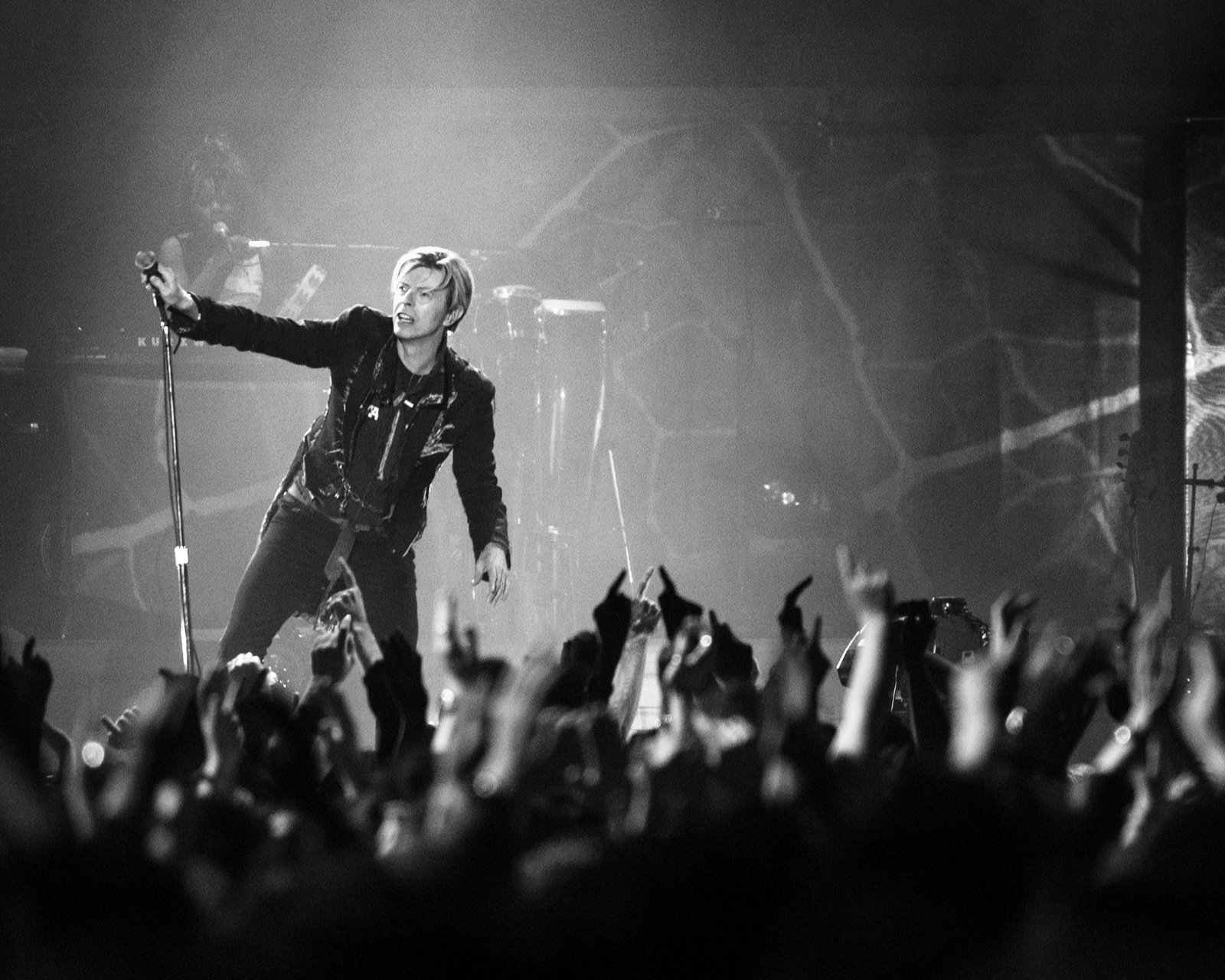 David Bowie singing on stage in a black jacket, reaching towards a crowd with raised hands, in black and white