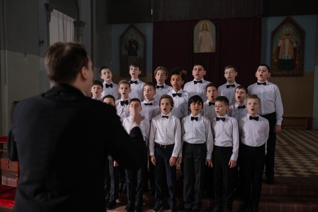 A boys' choir in white shirts and bow ties singing in a church while a conductor leads them