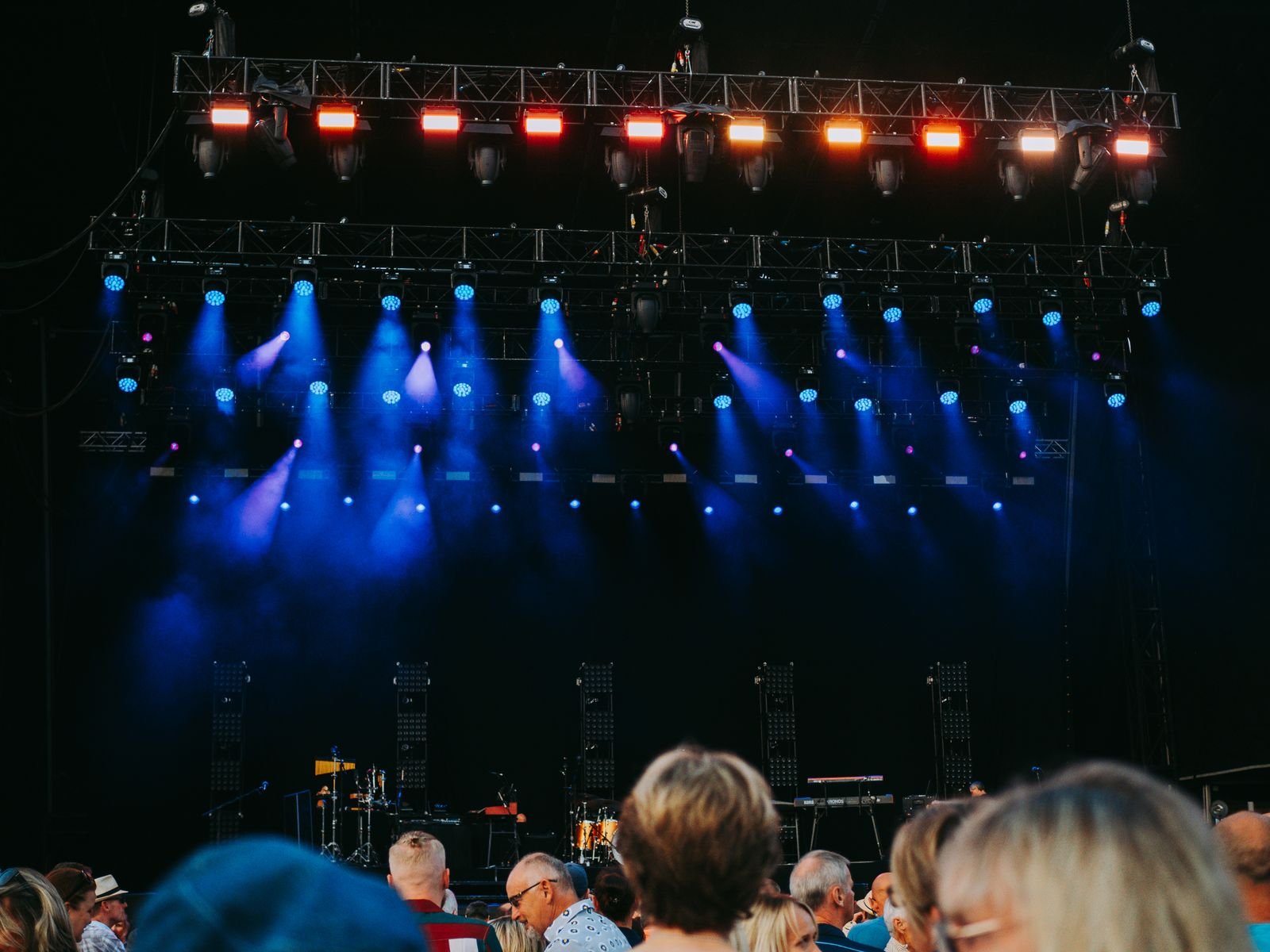 An outdoor concert audience in front of a stage lit by rows of blue and orange spotlights