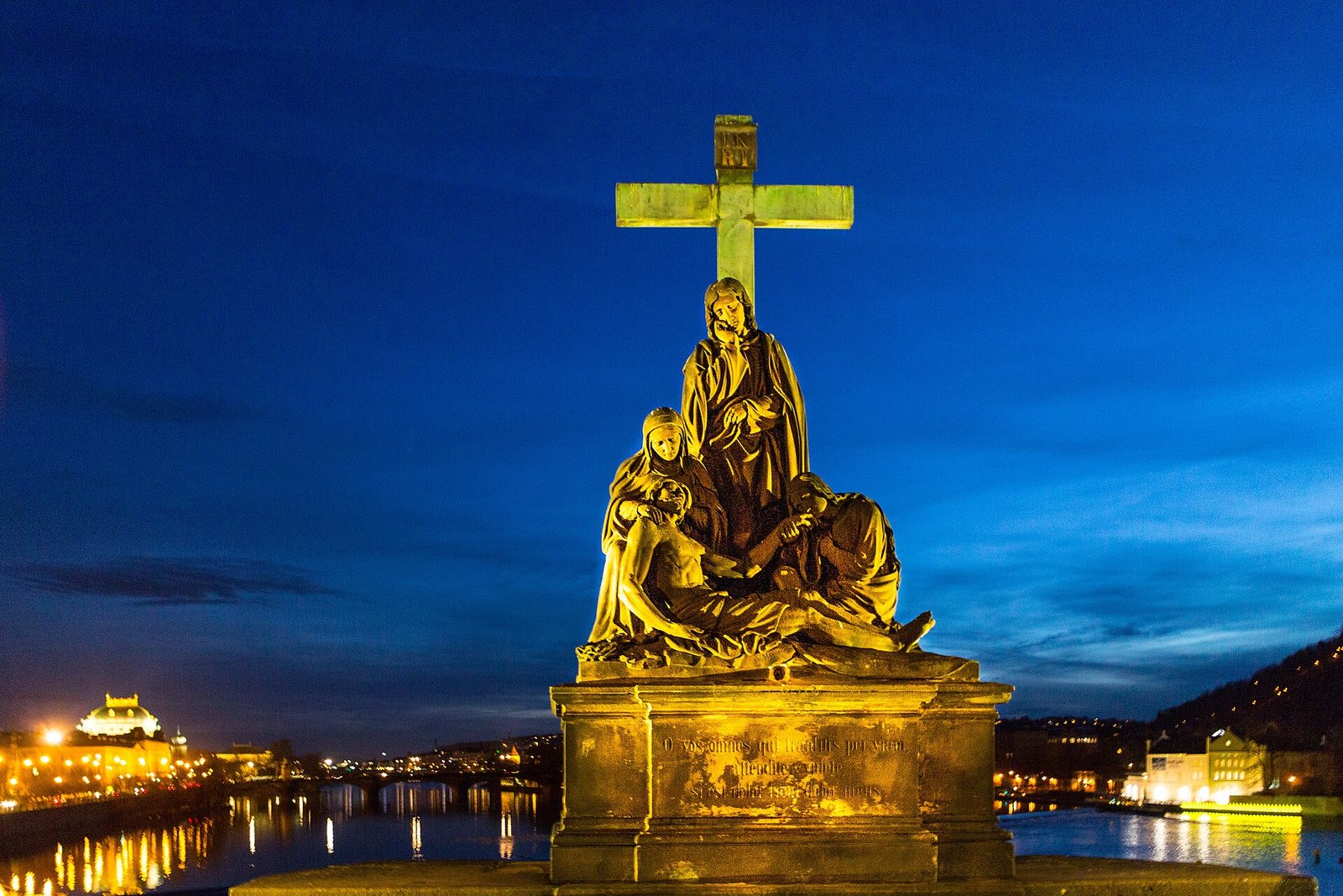 The Pieta sculpture on Prague's Charles Bridge lit at dusk, with the Vltava river and the National Theatre behind