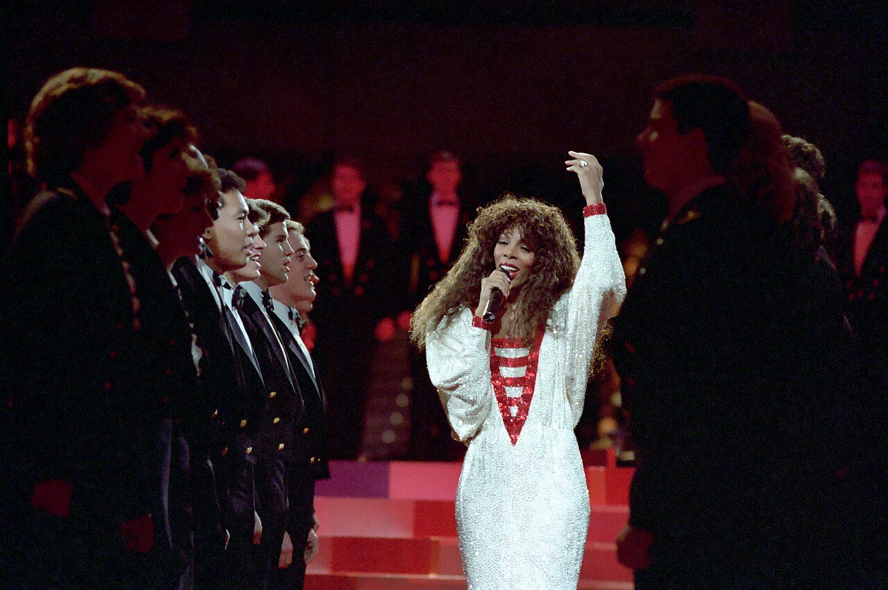 Donna Summer singing into a microphone on stage in a white and red sequined dress, backing singers in tuxedos behind her