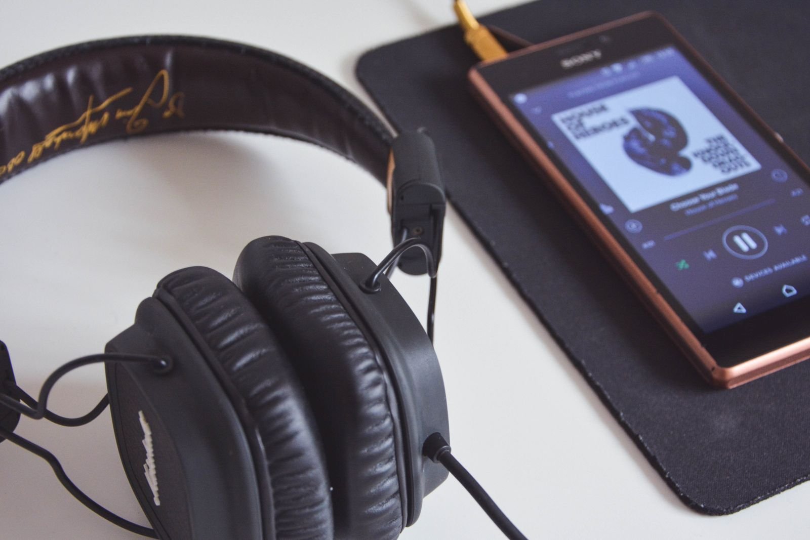 Black headphones on a white desk next to a smartphone playing a song in a music app