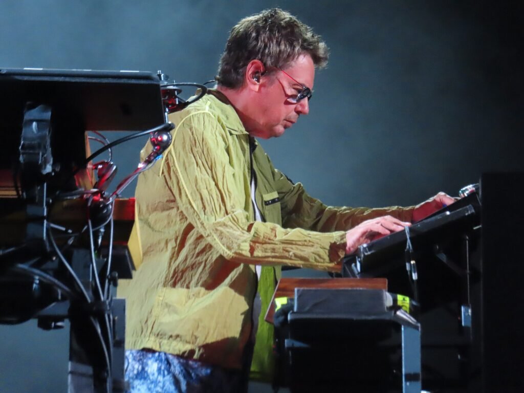 Jean-Michel Jarre in glasses and a yellow-green jacket plays a keyboard on a dark stage
