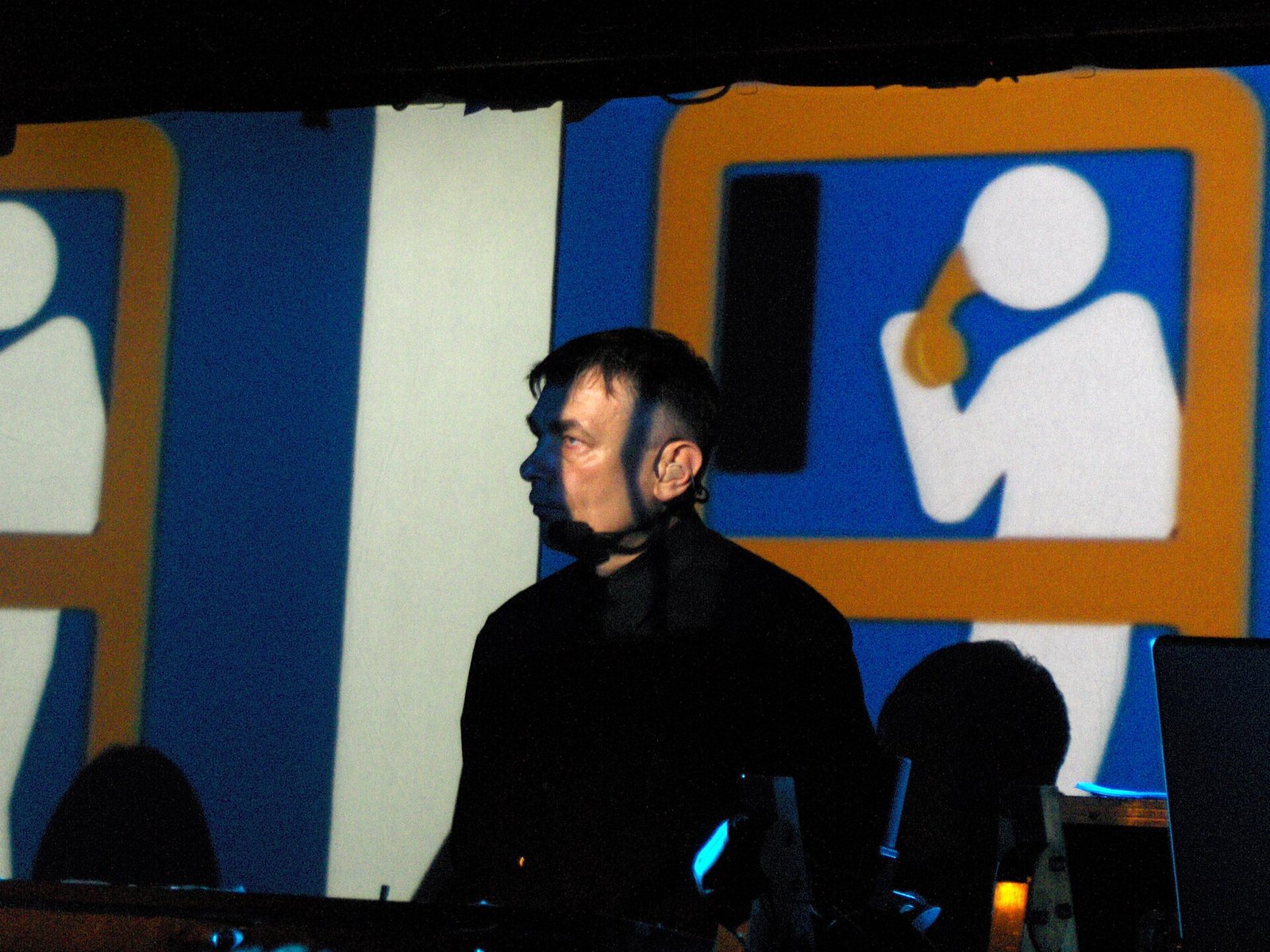 Karl Bartos in a black shirt on a dark stage in front of a projected blue and orange graphic of a figure holding a telephone