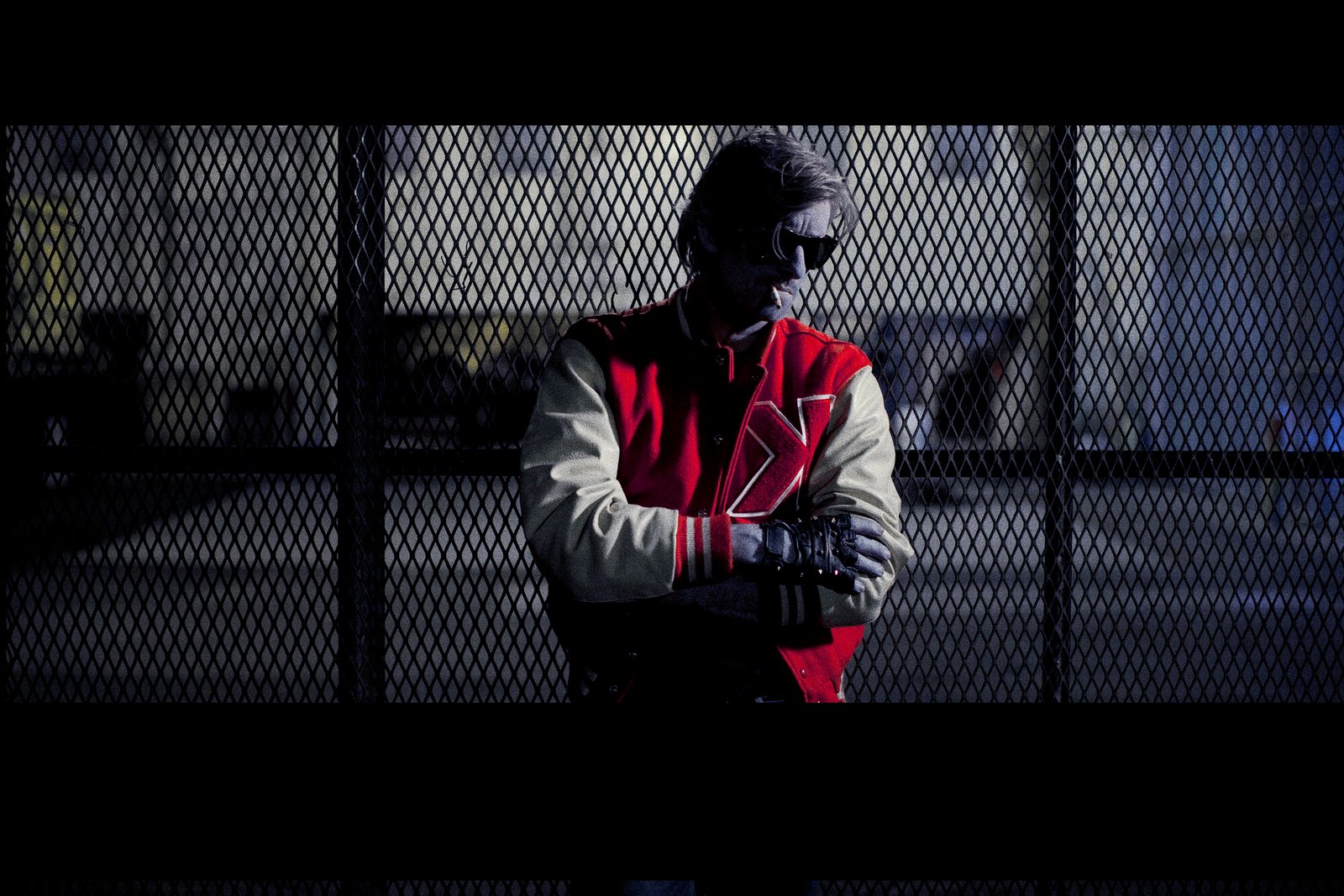 Kavinsky in his red and white varsity jacket and sunglasses, photographed through a chain-link fence
