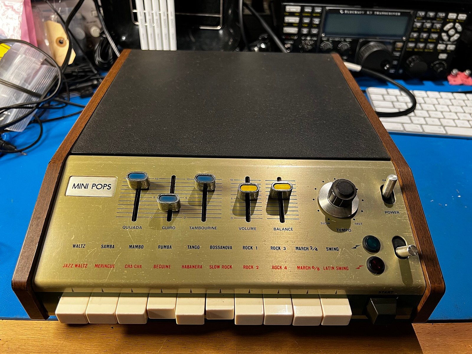 A Korg Mini Pops 7 rhythm box with wooden side panels, three percussion sliders and two rows of rhythm names above white piano-style keys