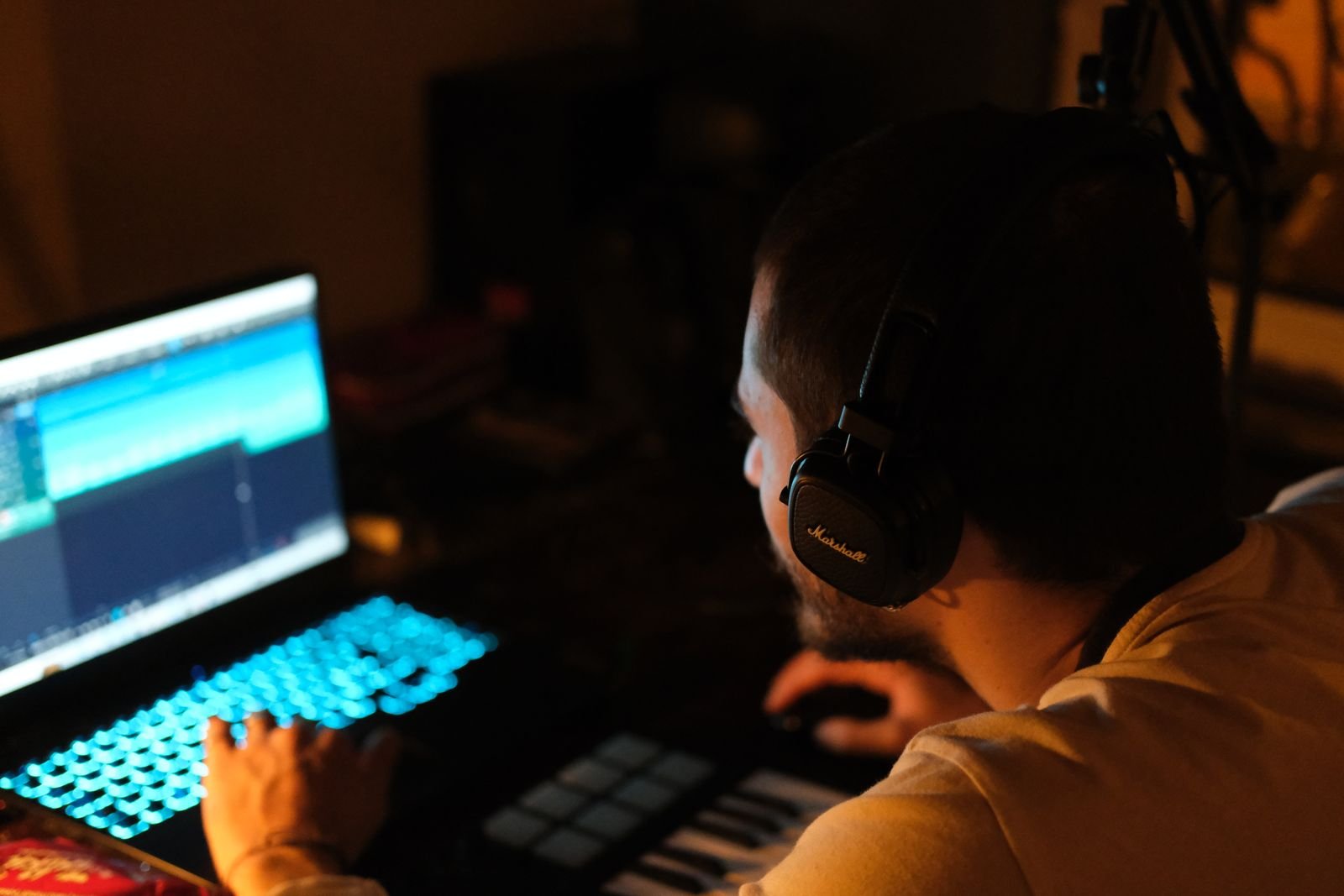 A man in headphones works on a music track on a laptop next to a MIDI keyboard in a dim room