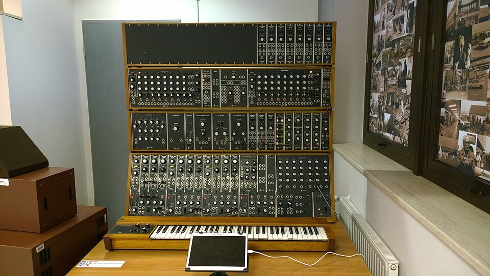 A large 1970s Moog modular synthesizer with rows of knobs and patch cables above a wooden keyboard