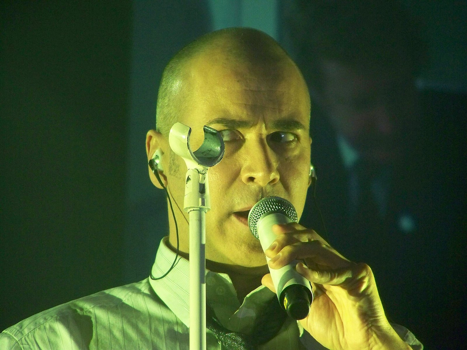 Philip Oakey of the Human League singing into a handheld microphone under green stage light