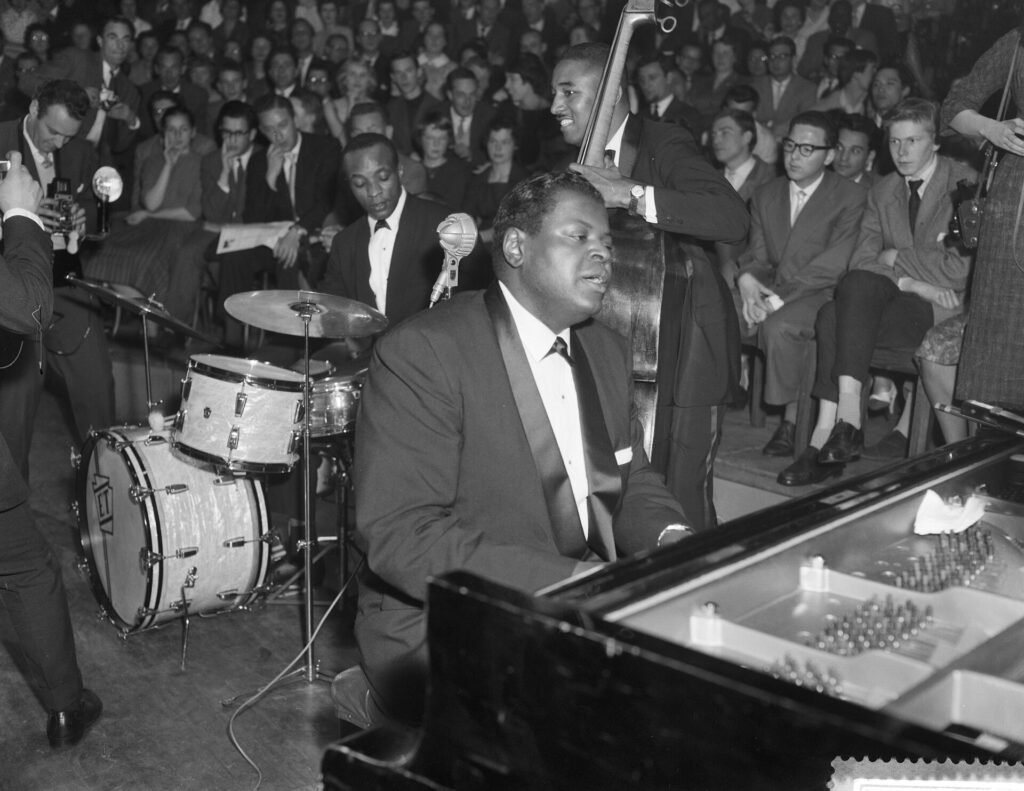 Oscar Peterson playing a grand piano on stage with bassist Ray Brown and drummer Ed Thigpen behind him, in black and white