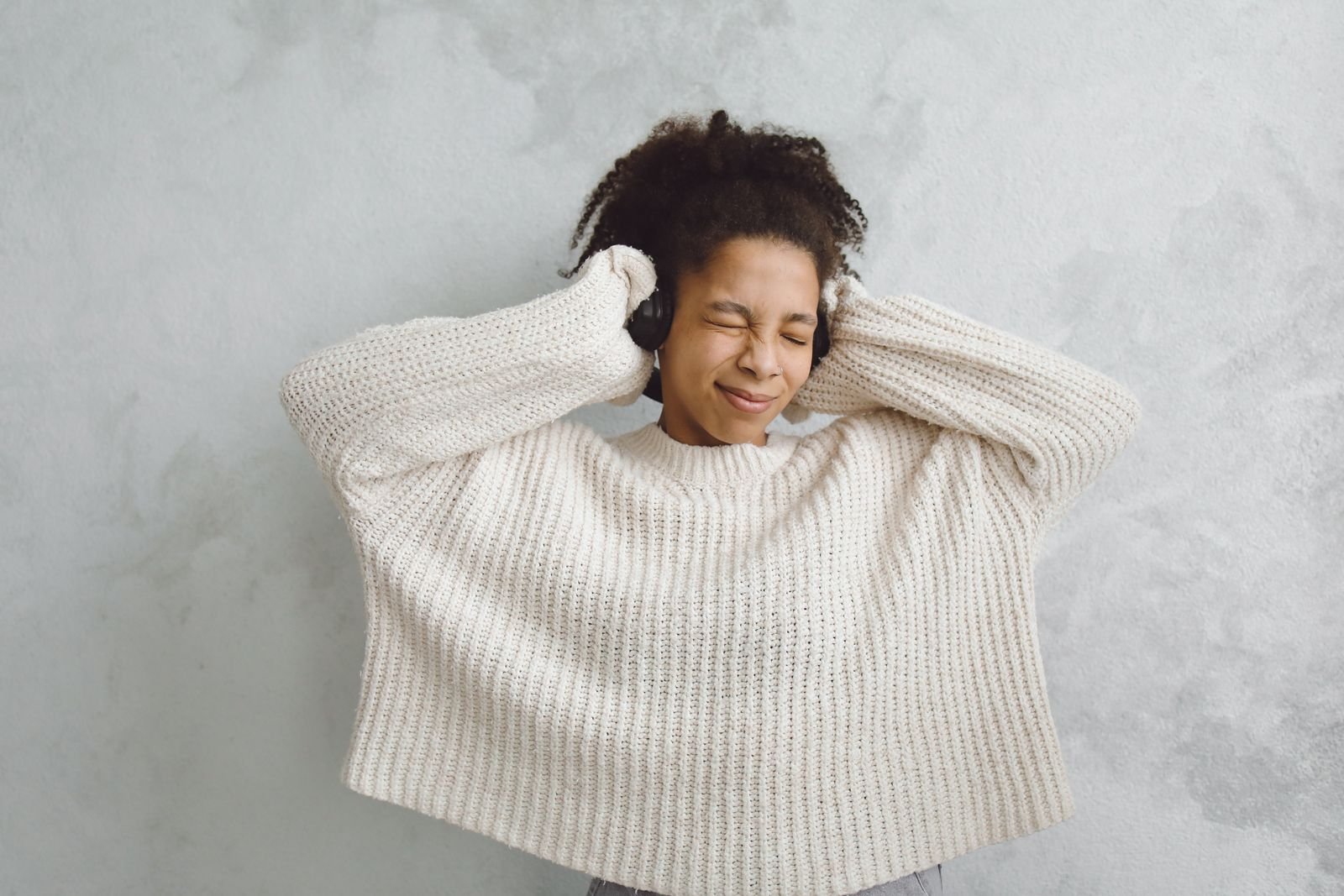 A woman with curly hair and eyes closed holds her headphones while listening to music