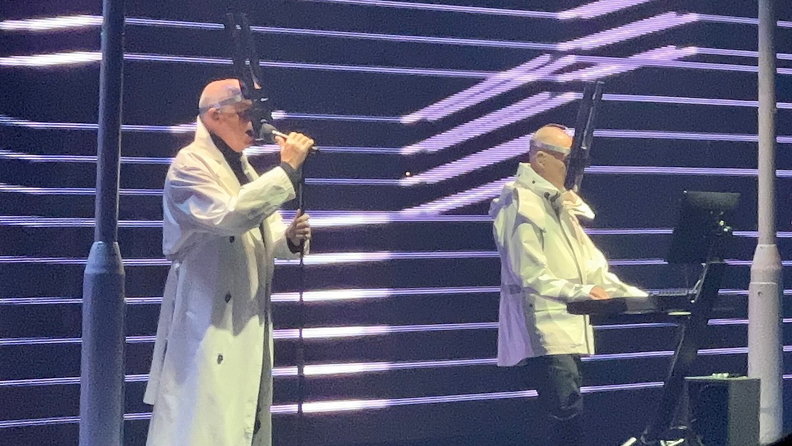 Neil Tennant singing in a white coat and Chris Lowe at a keyboard on a stage lit with white light lines